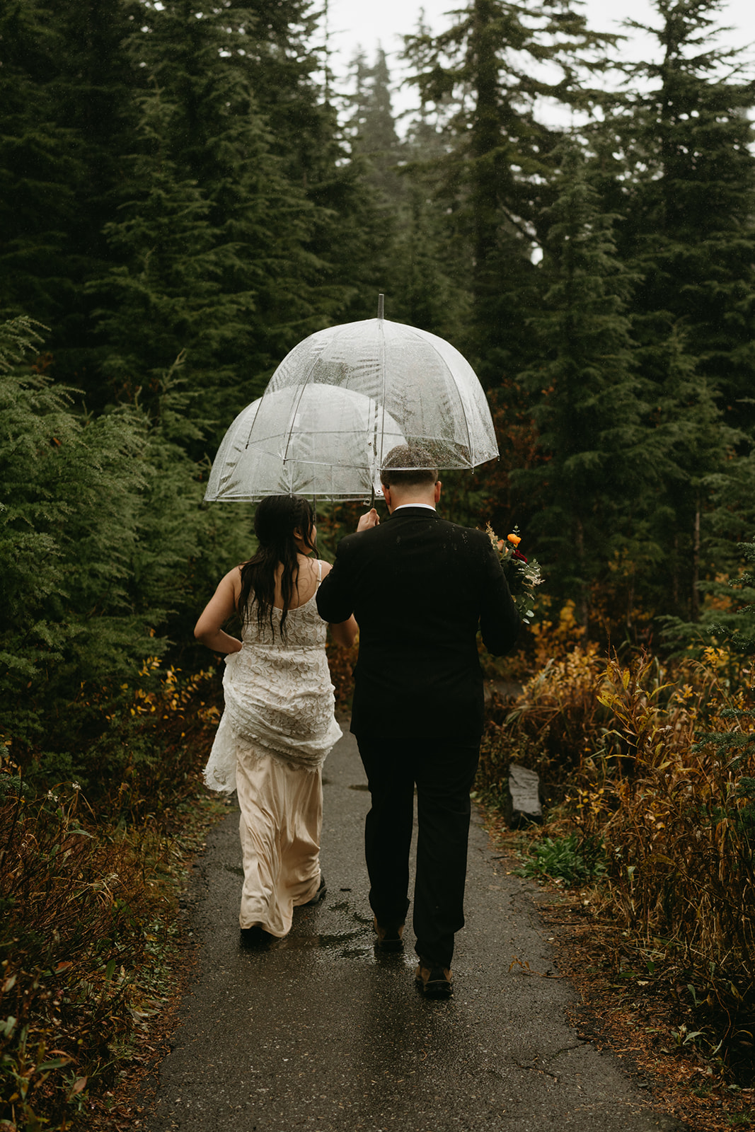 Couple walking hand in hand through the forest trail under umbrellas, soaking in their rainy wedding.