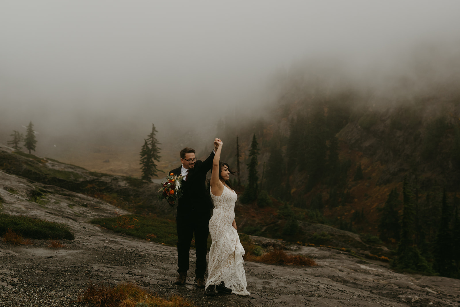 Groom twirling the bride as they dance on a foggy mountain ledge during their rainy wedding.