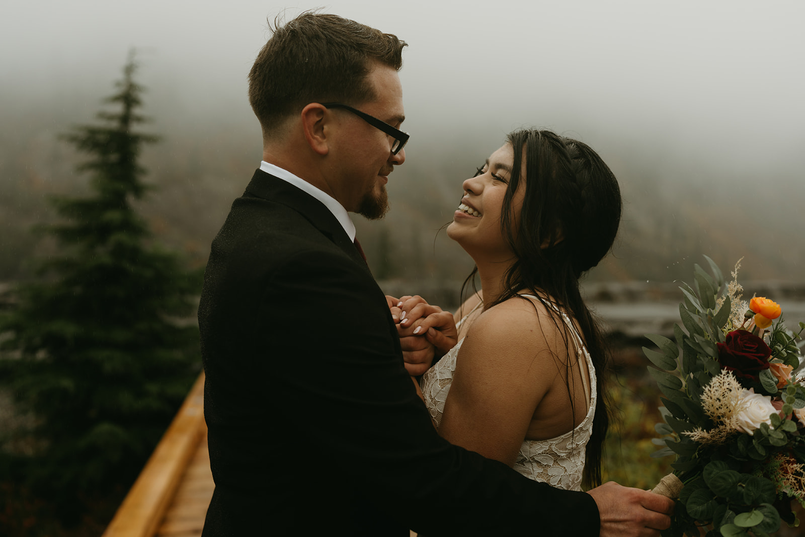 Joyful bride and groom laughing and holding hands with fog and forest behind them.