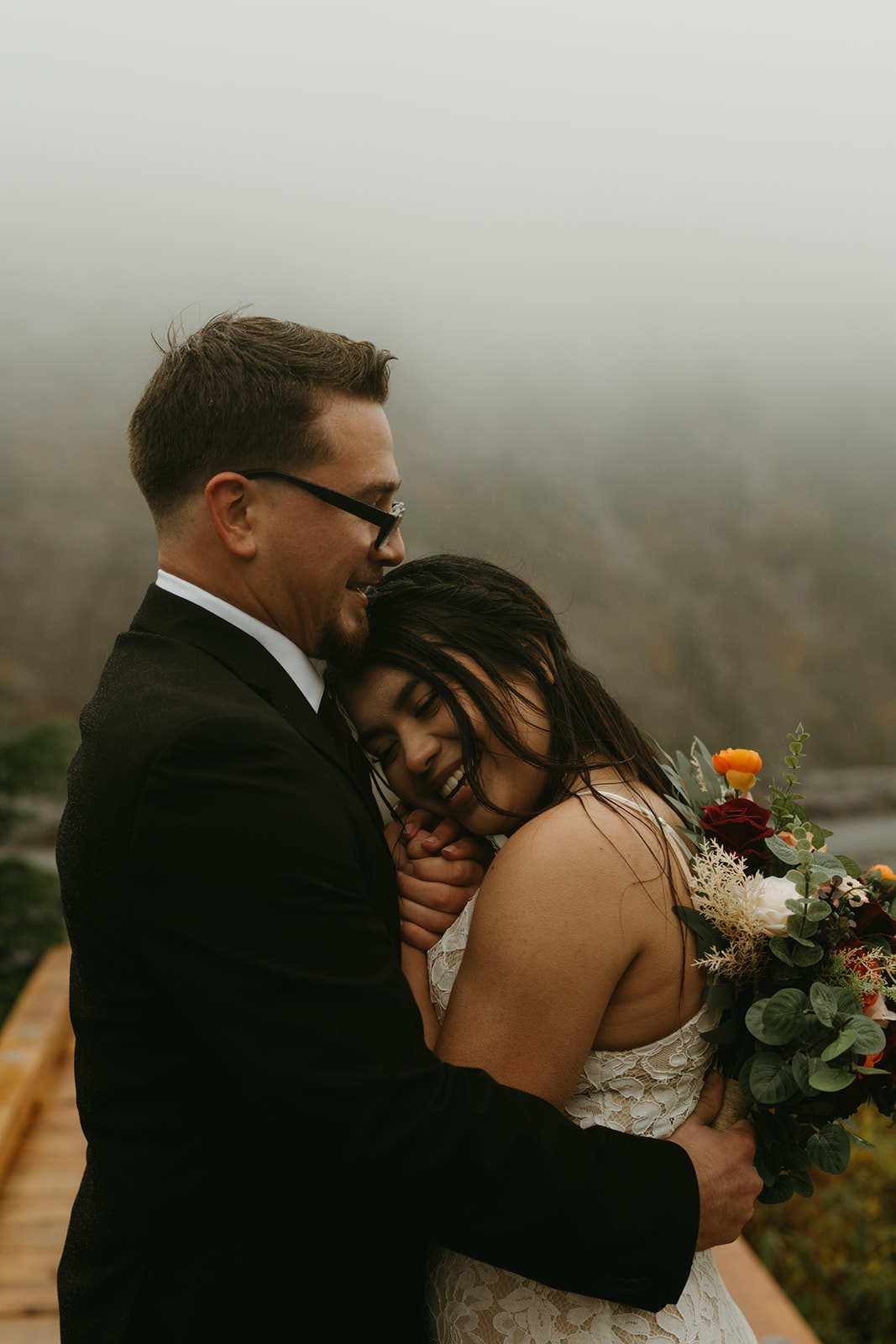 Bride resting her head on the groom’s chest, holding a colorful bouquet on a misty mountain path.