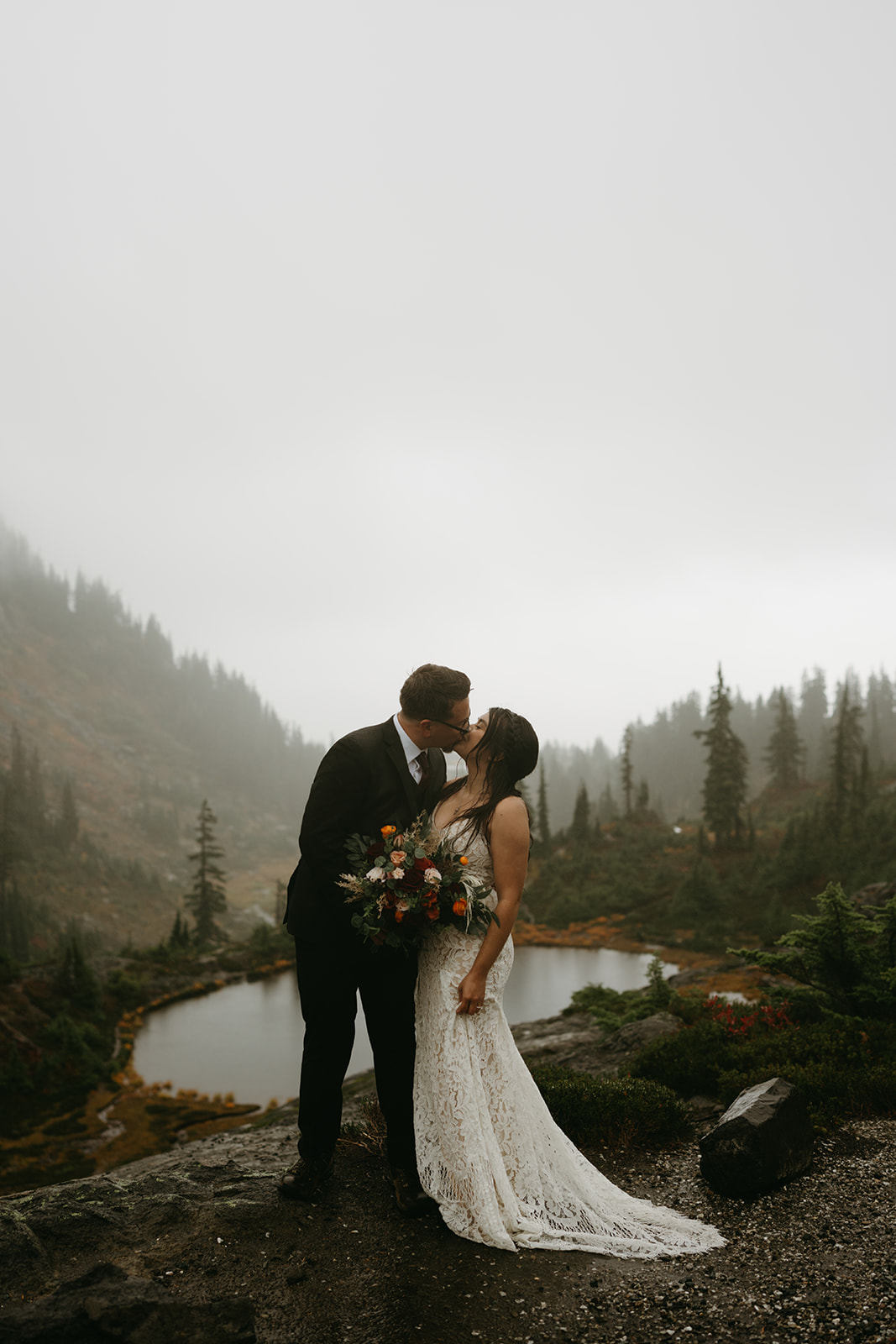 Bride and groom kissing in front of a misty alpine lake surrounded by fall colors.