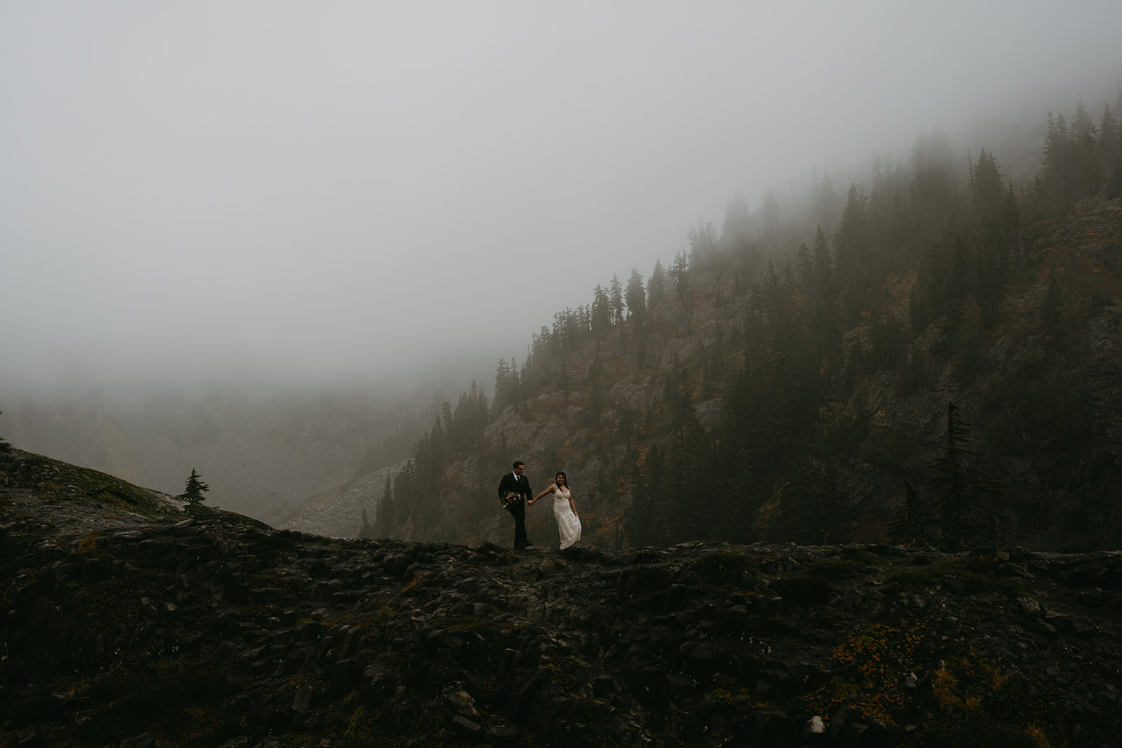 Bride and groom walk along a dramatic, fog-covered ridge during their adventurous rainy wedding.