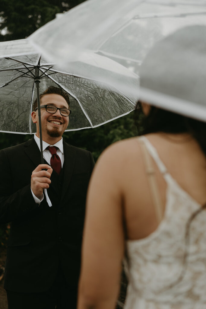 Groom smiling warmly at his bride under an umbrella during their rainy wedding vows.