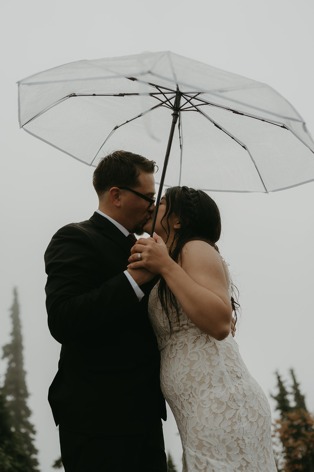 Bride and groom share a kiss under a clear umbrella during their intimate rainy wedding ceremony.