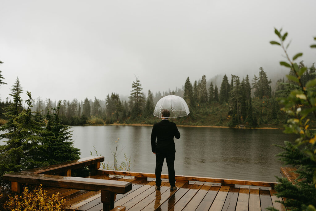Groom overlooking the foggy lake, standing alone with an umbrella during his rainy wedding.