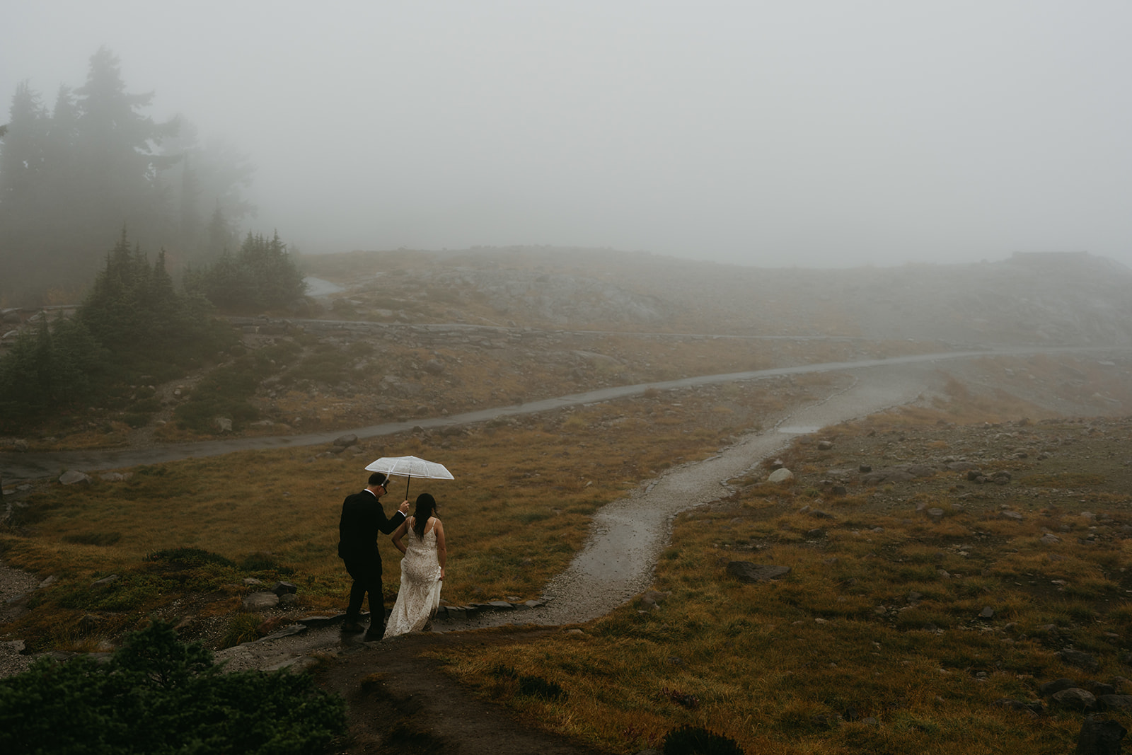 Couple walking into the fog, sharing an umbrella during their quiet, romantic rainy wedding.