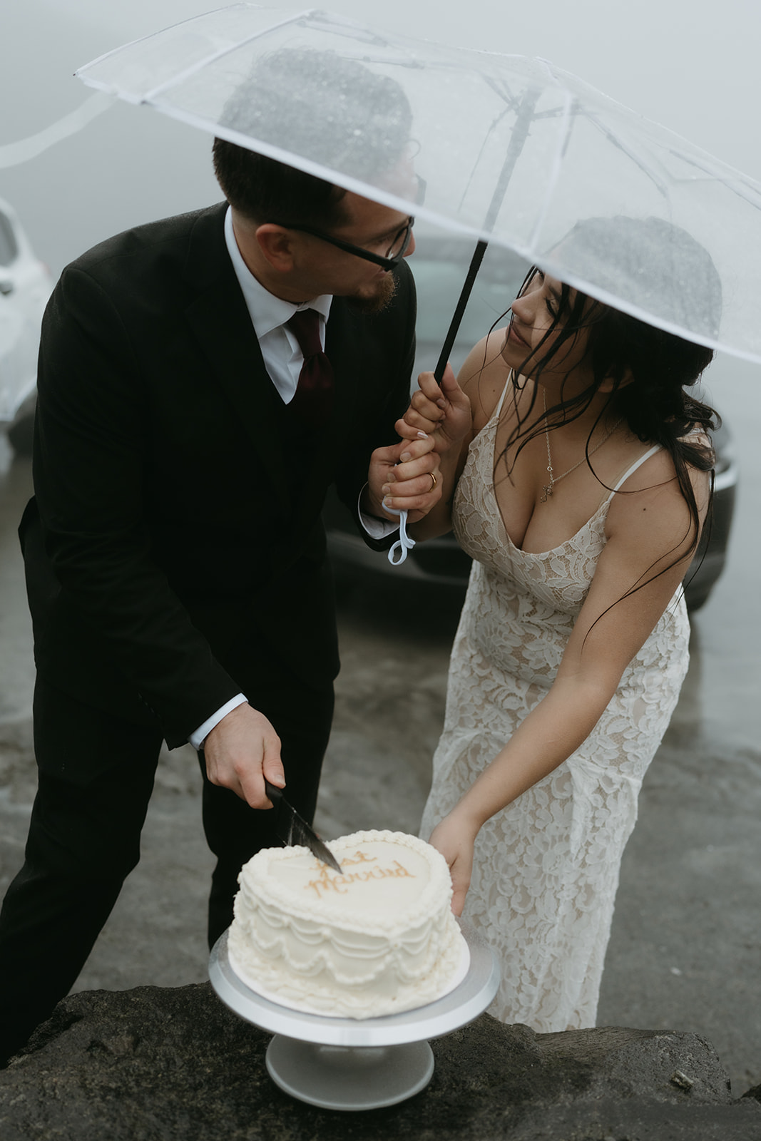 Couple cutting their wedding cake together under an umbrella during their rainy wedding.