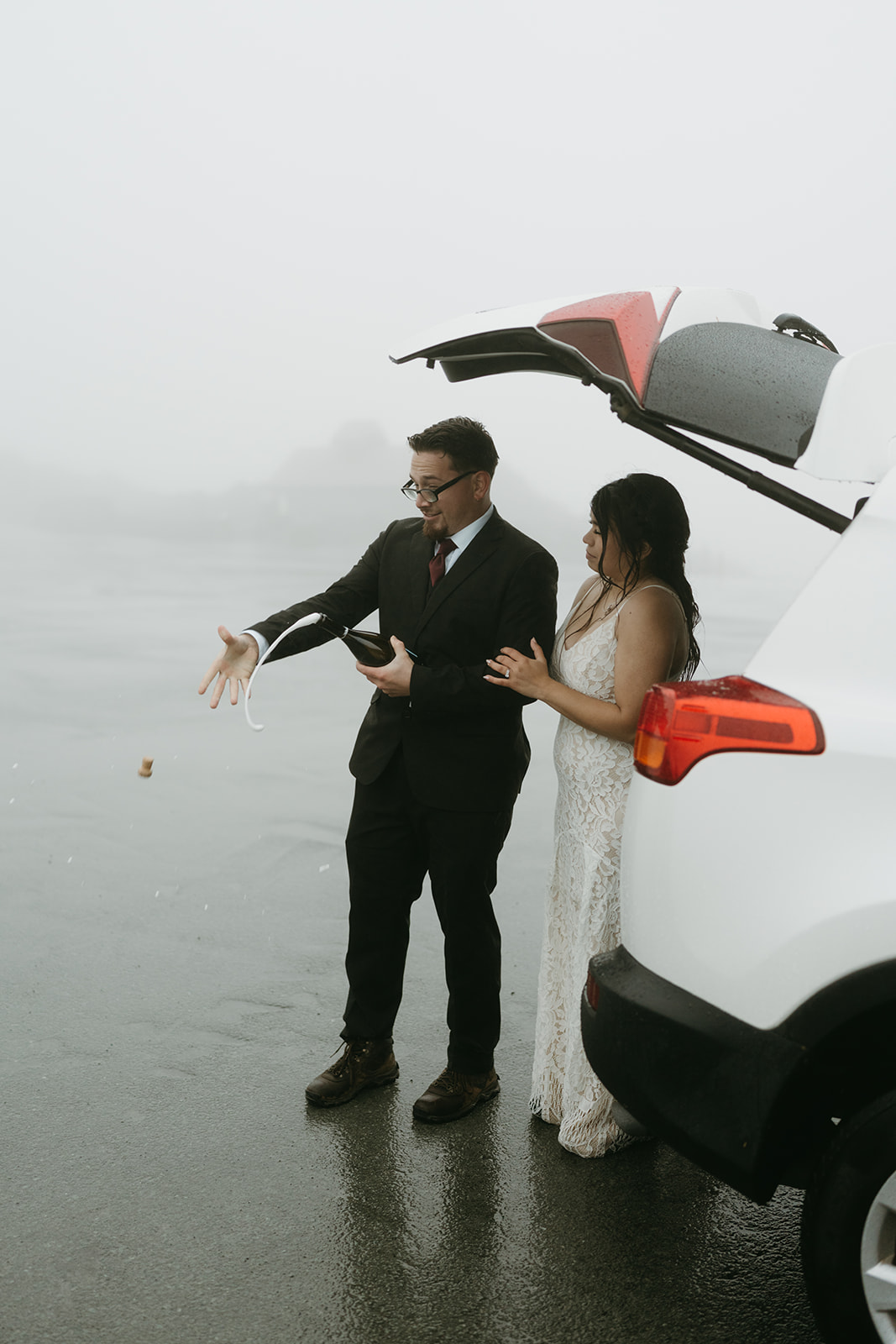 Bride and groom popping champagne by their car after their foggy mountain rainy wedding.