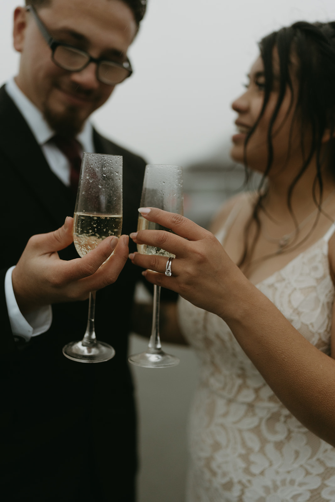 Close-up of champagne flutes clinking with wedding rings in focus during a rainy celebration.