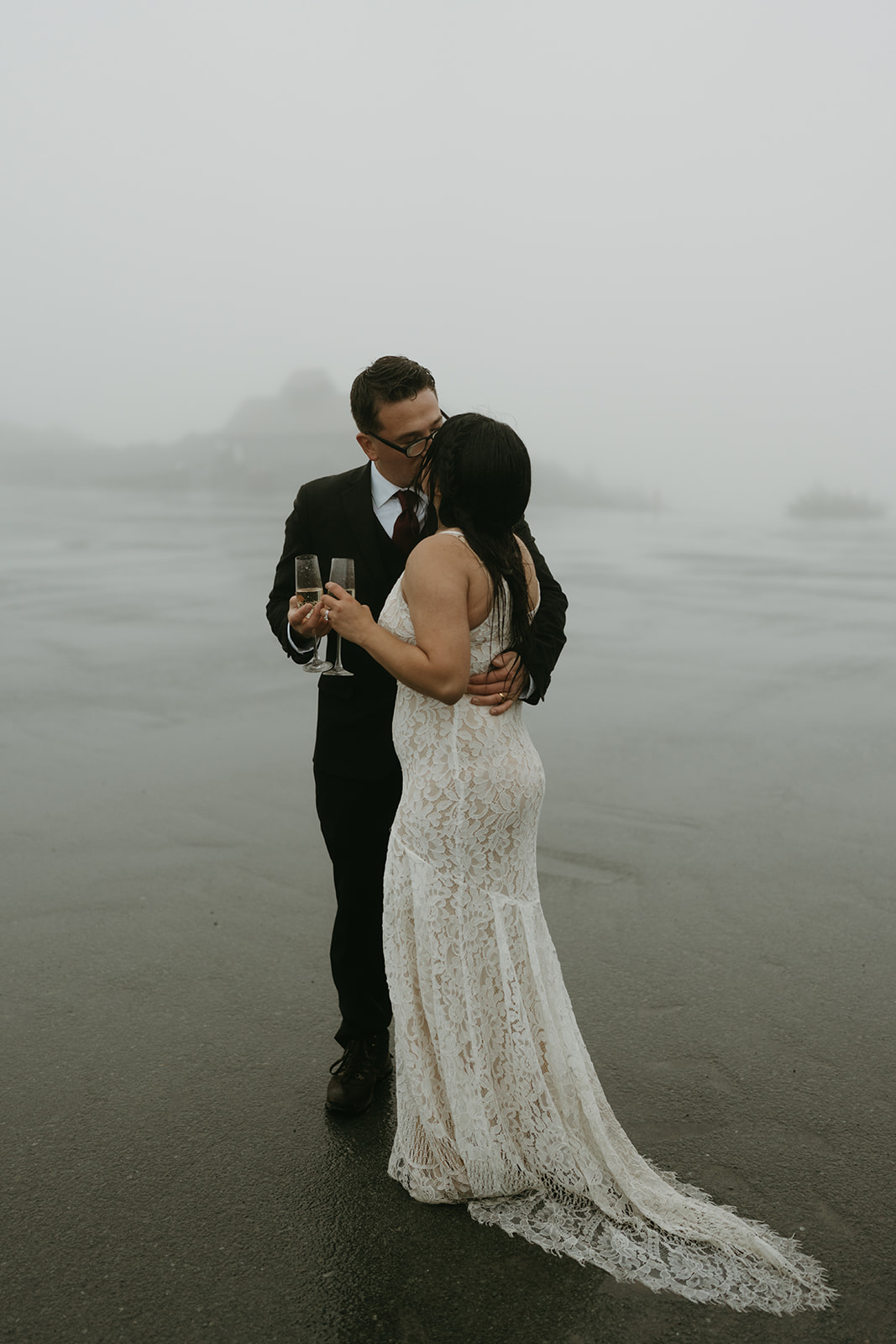 Bride and groom share a kiss with champagne in hand, surrounded by fog and quiet joy.