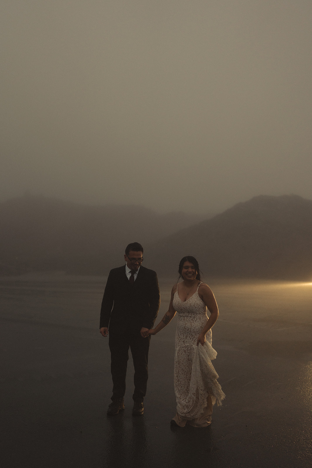 Bride and groom walking hand in hand through the mist, smiling in the soft light.