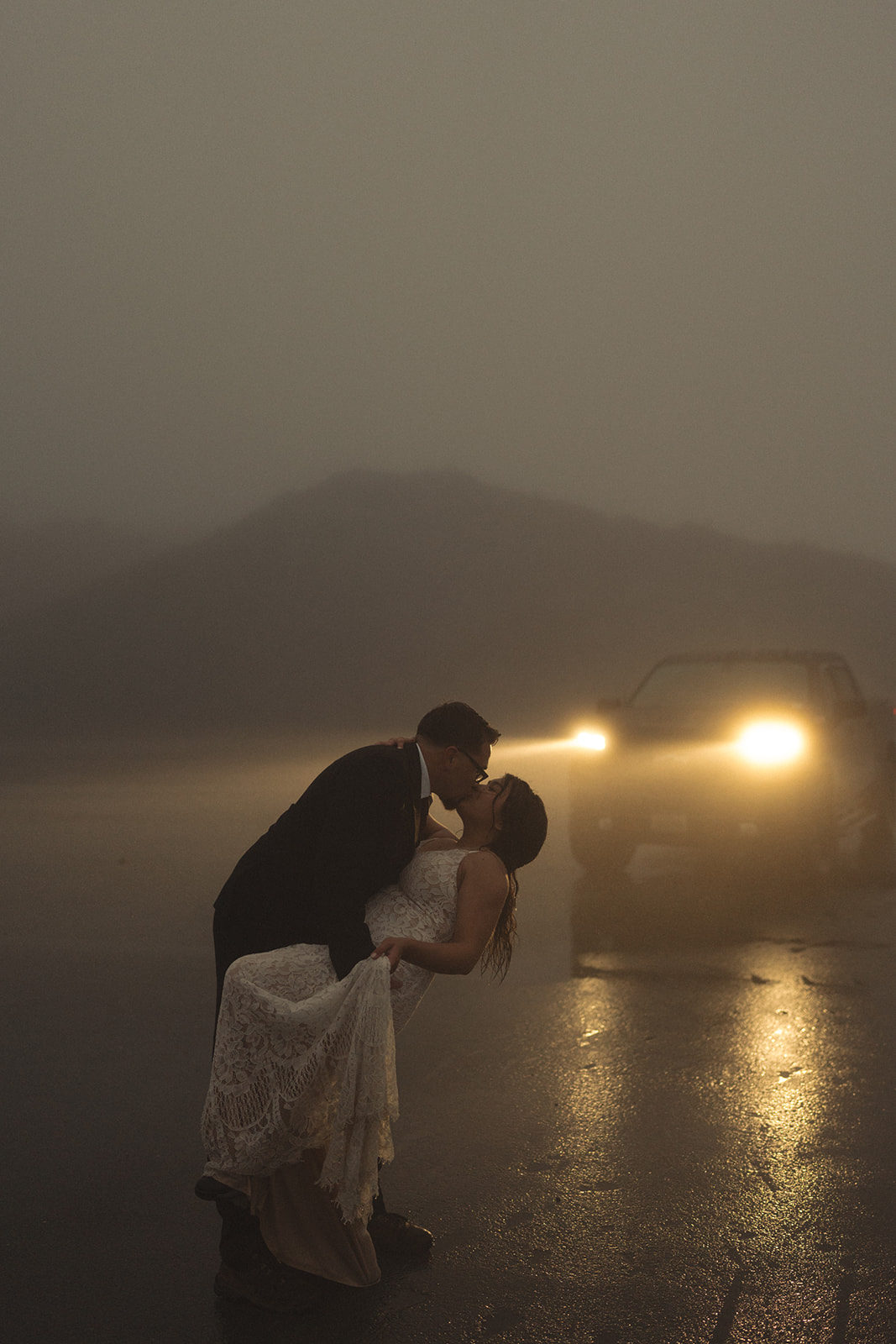 Groom dipping the bride for a kiss in the rain, headlights glowing behind them during a rainy wedding.