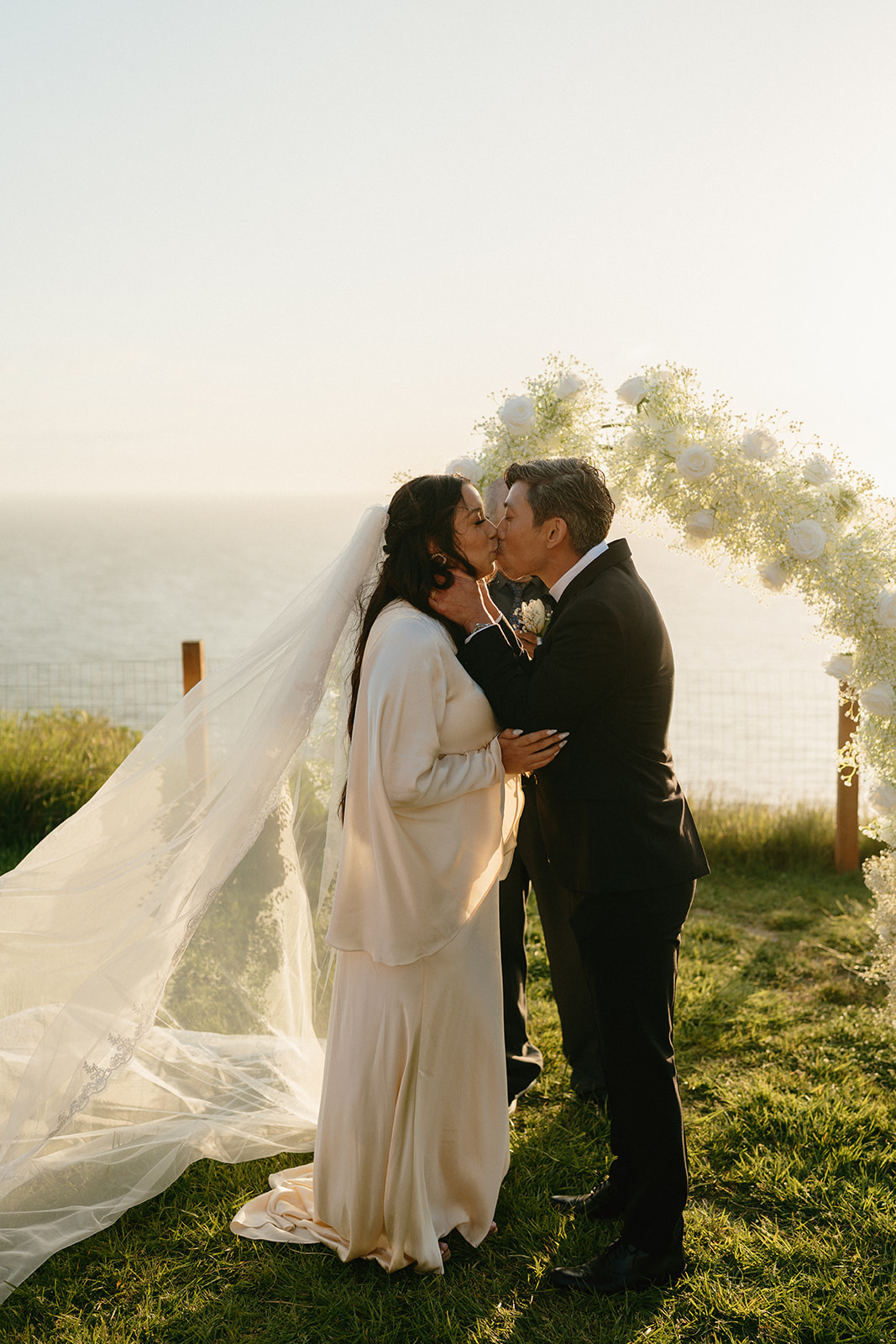 The couple shares their first kiss beneath a floral arch overlooking the sea, surrounded by golden light.
