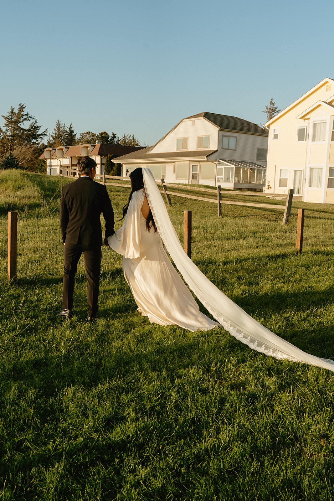 Bride and groom walking hand in hand toward a quiet inn, her long veil trailing in the grass.