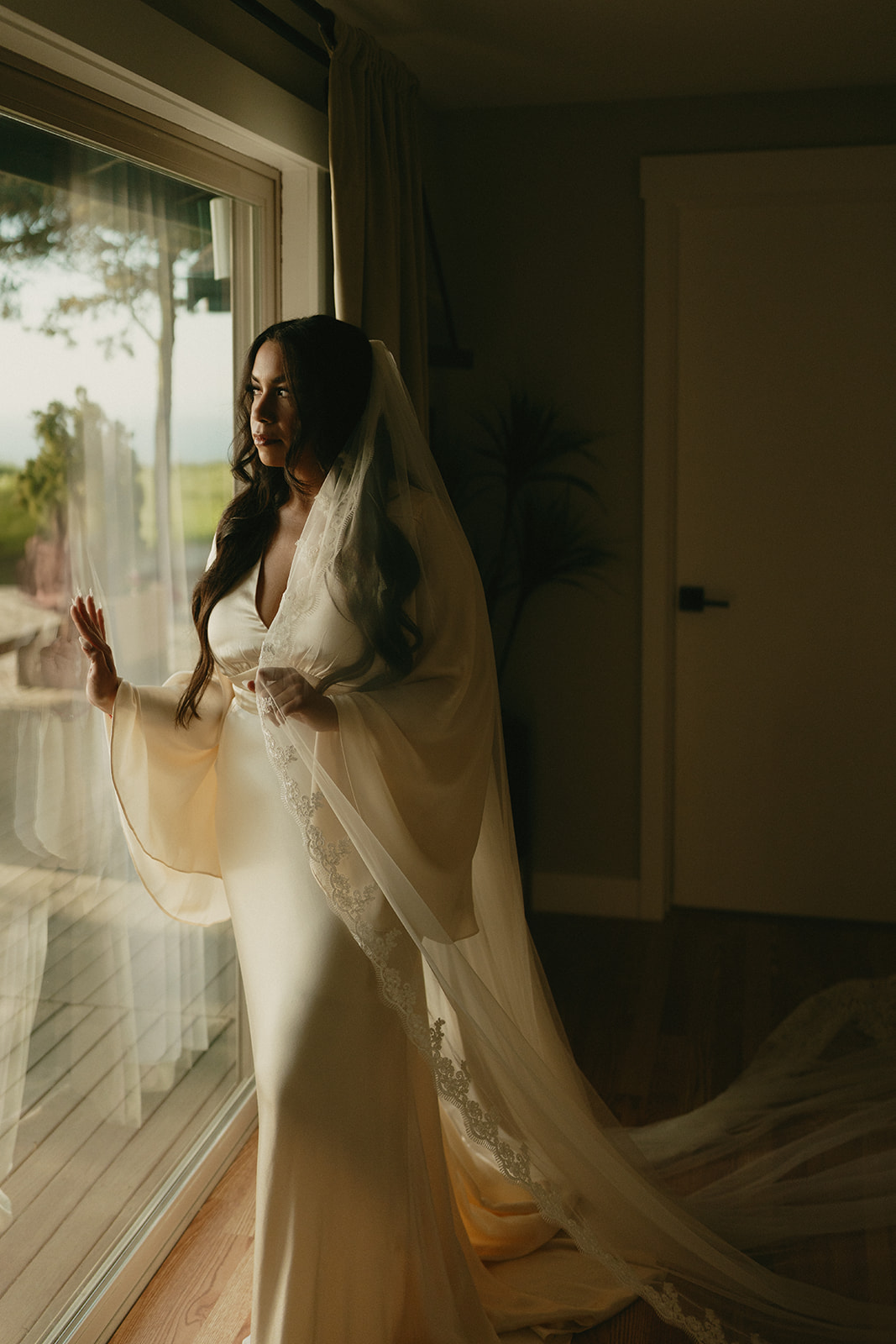 Bride standing by a window in an elegant satin gown and veil, gazing out at the view on Whidbey Island.