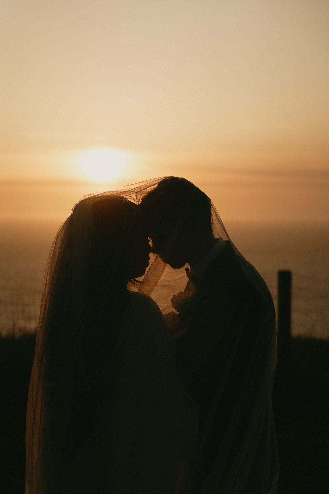 Silhouette of the couple sharing a tender moment under the bride’s veil as the sun sets behind them.