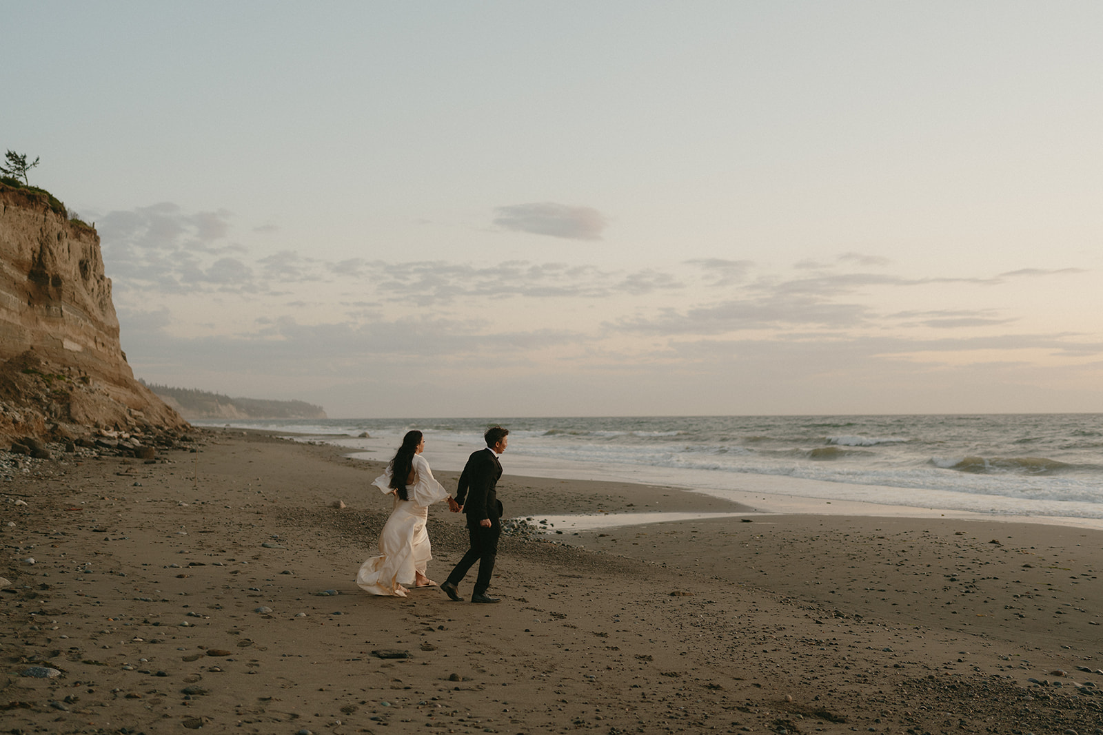 Romantic beach walk along the shore, the couple framed by sea cliffs and the soft dusk sky on Whidbey Island. 
