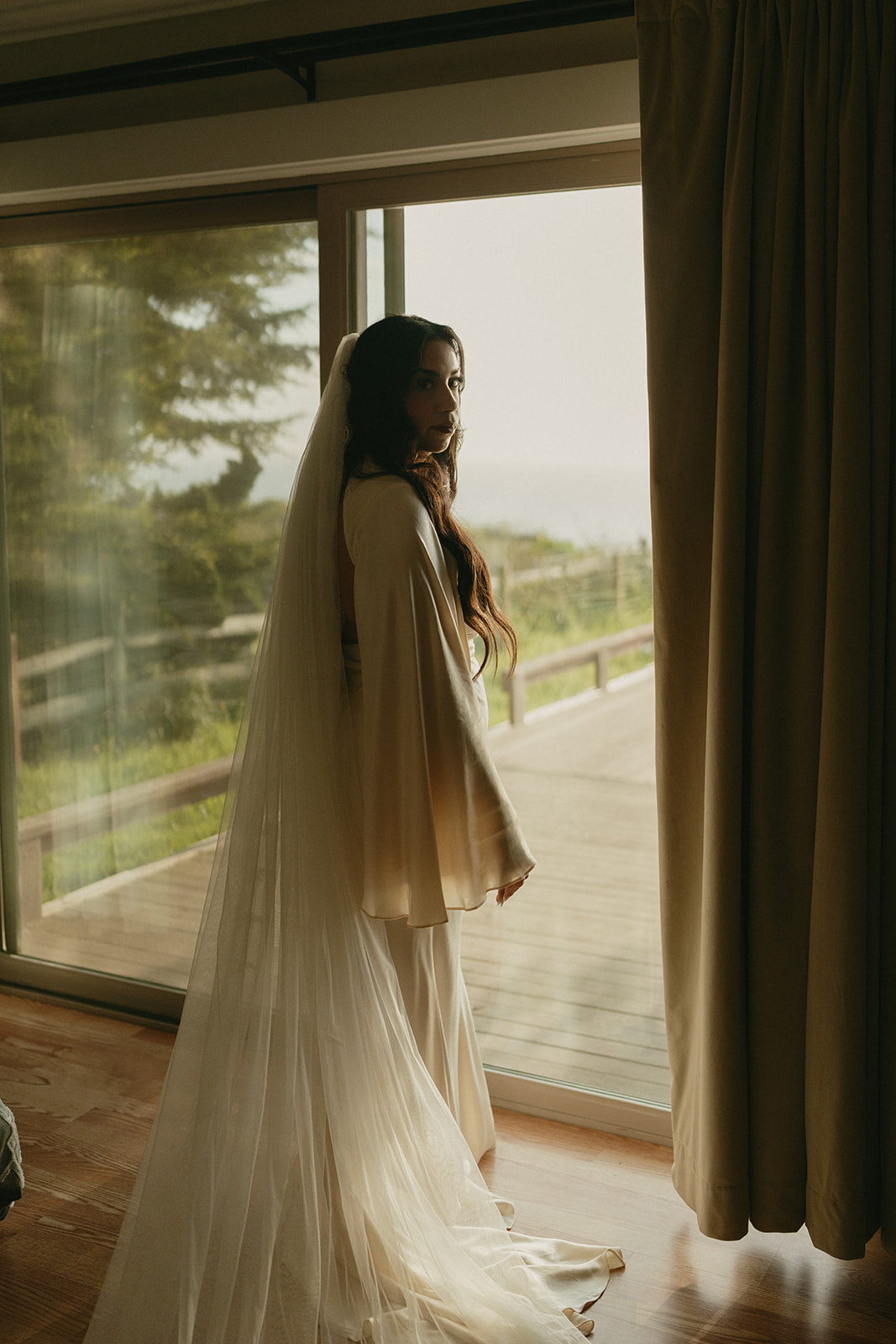 Bride looking over her shoulder as she stands by the glass door, light pouring into the room.