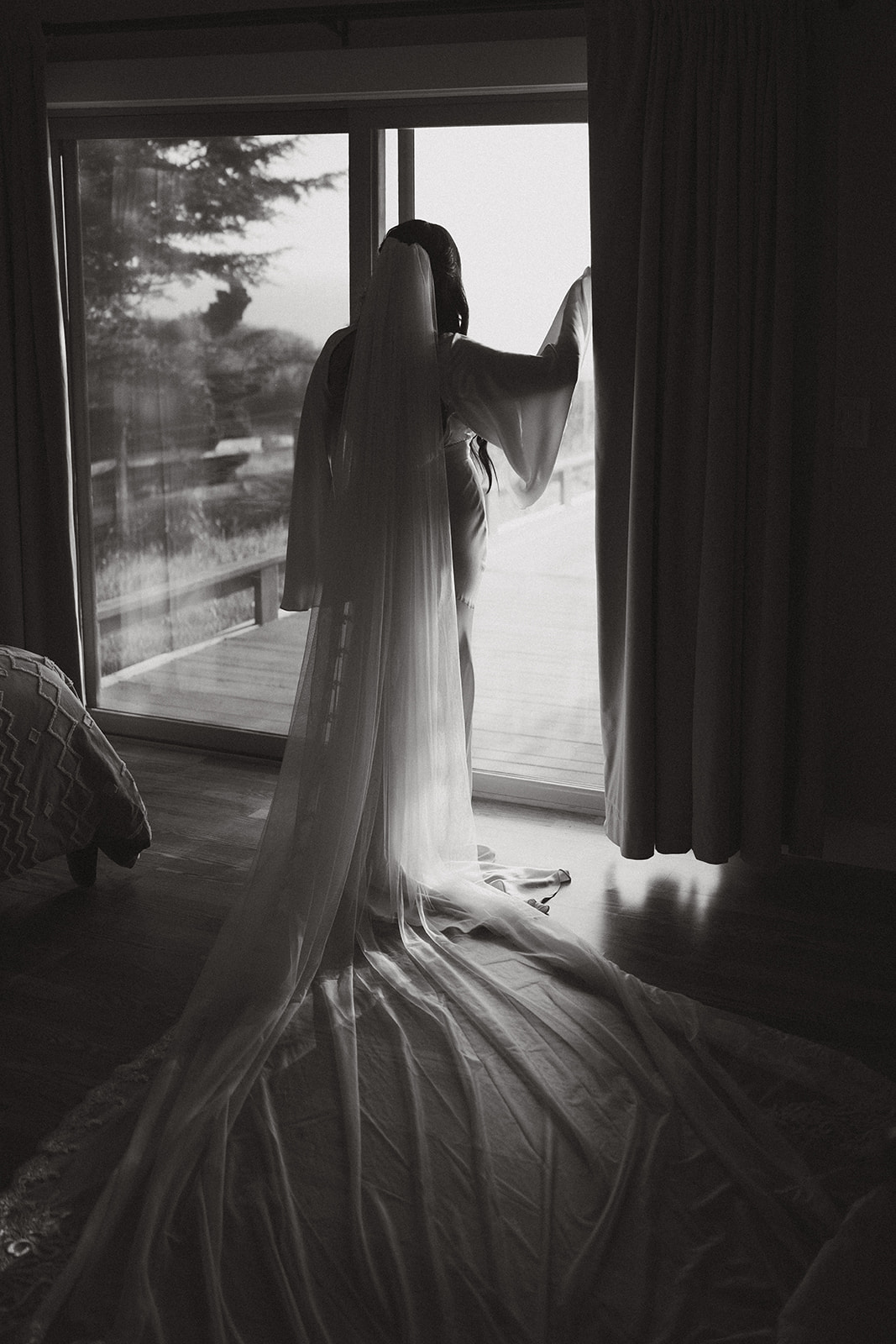Black and white photo of the bride standing by a window, her long veil spread across the floor.