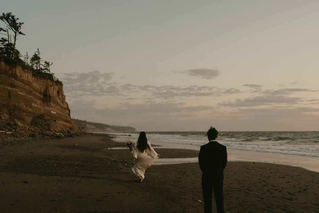 Bride barefoot in the sand while the groom looks on, the coastline stretching into the distance.