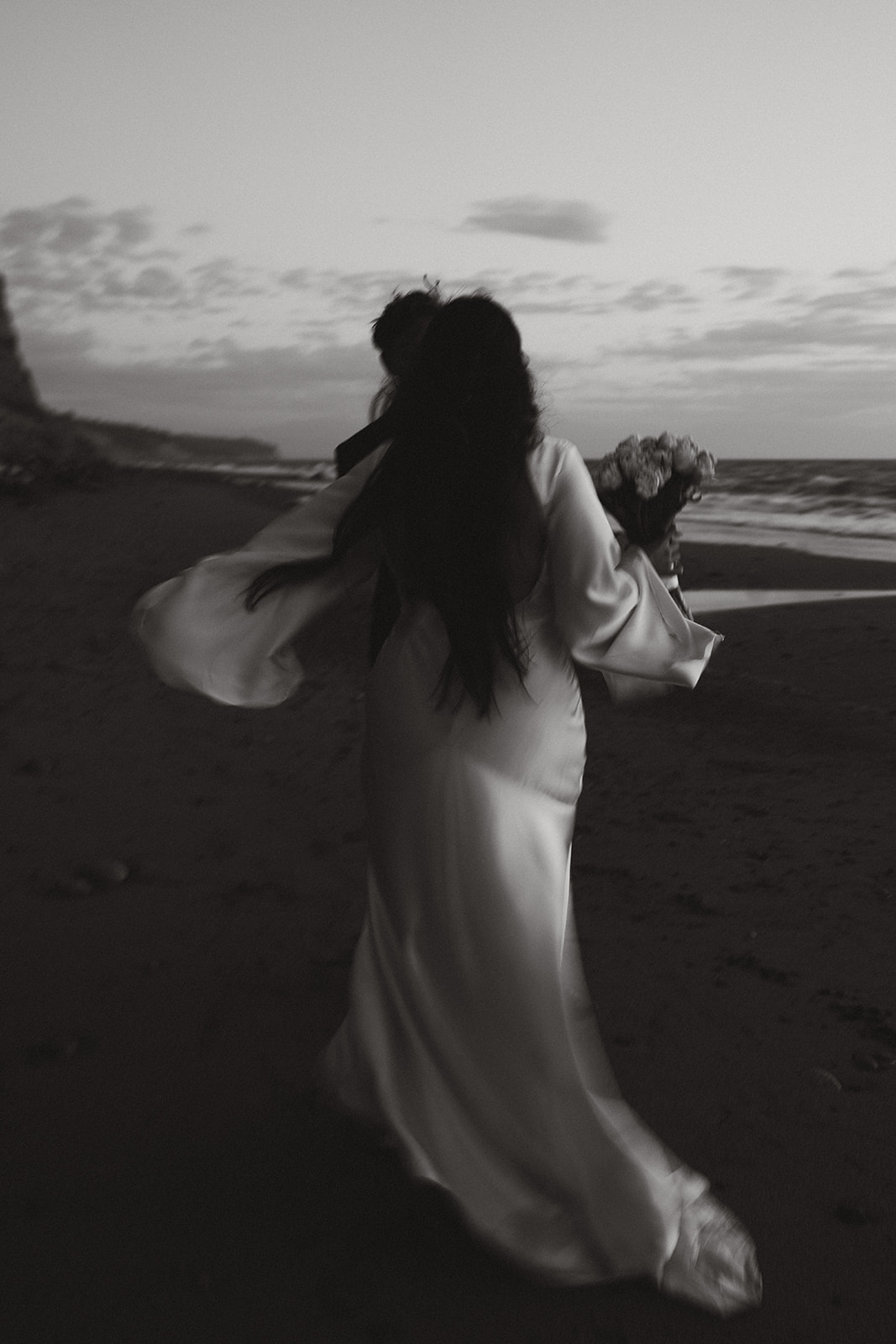 Black and white image of the couple walking down the beach, bride’s dress flowing behind her.