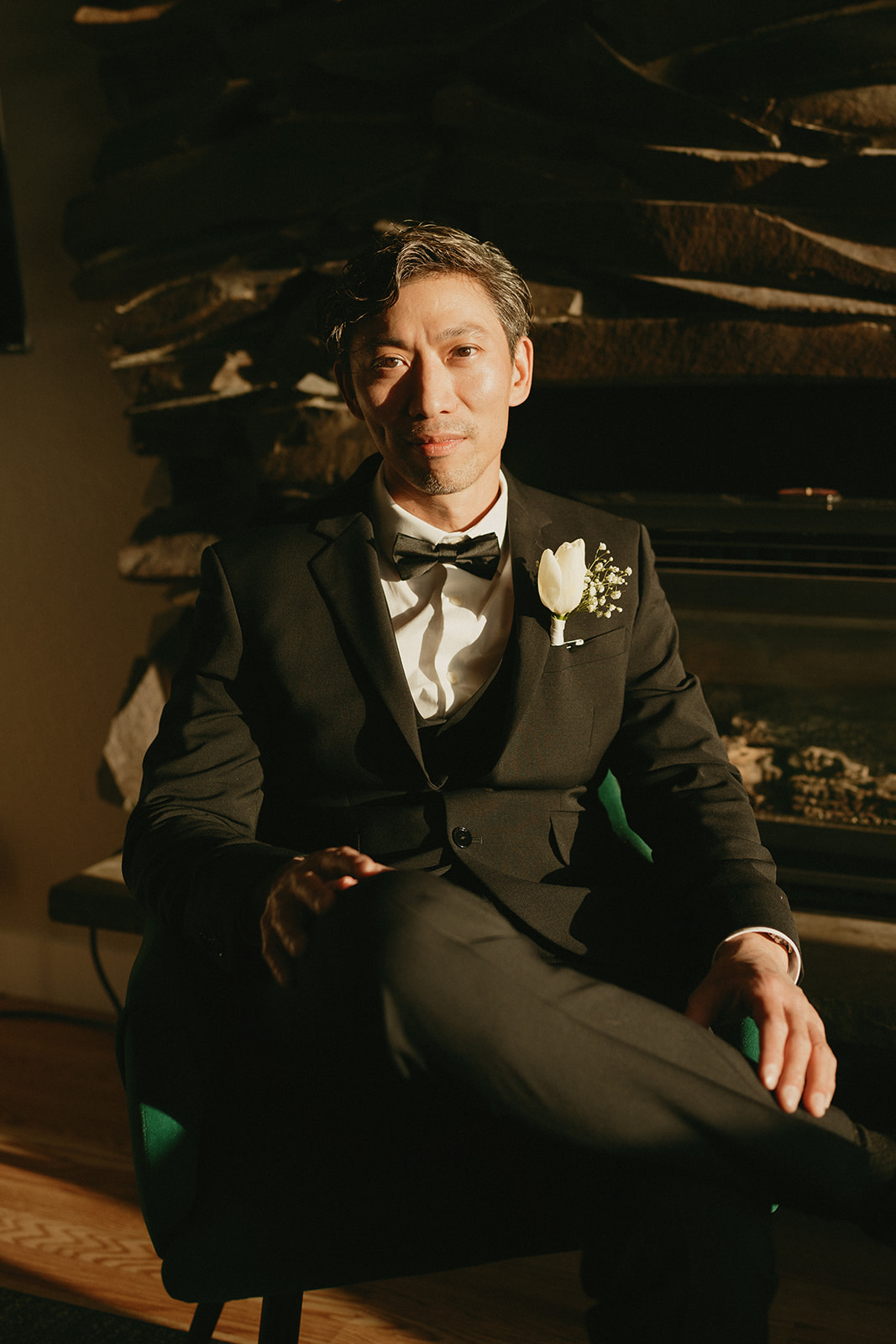 Groom in soft golden hour light, seated in a black suit with a white boutonniere near the fireplace.