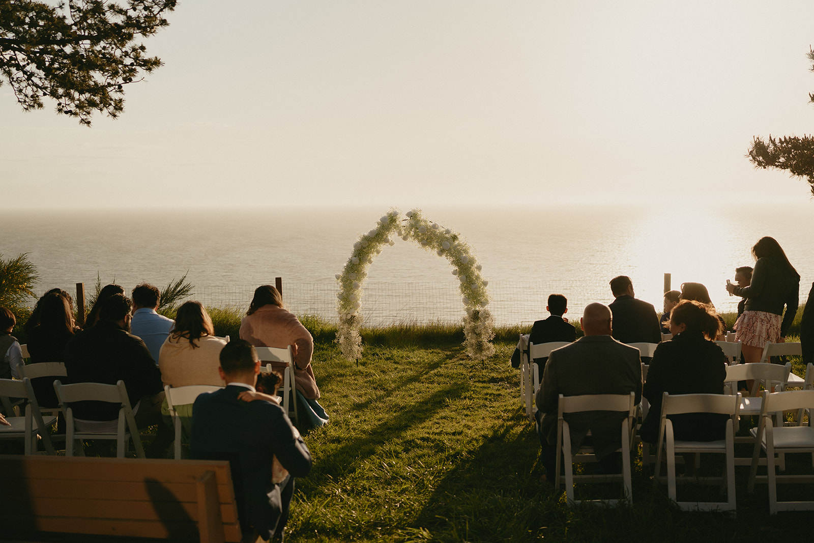 Guests seated and facing the ocean as the ceremony begins, the floral arch glowing in the late afternoon light.