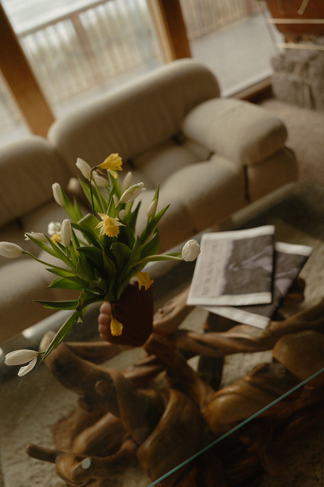 A bouquet of white and yellow tulips rests on a driftwood glass coffee table, next to a folded newspaper in a quiet coastal home.