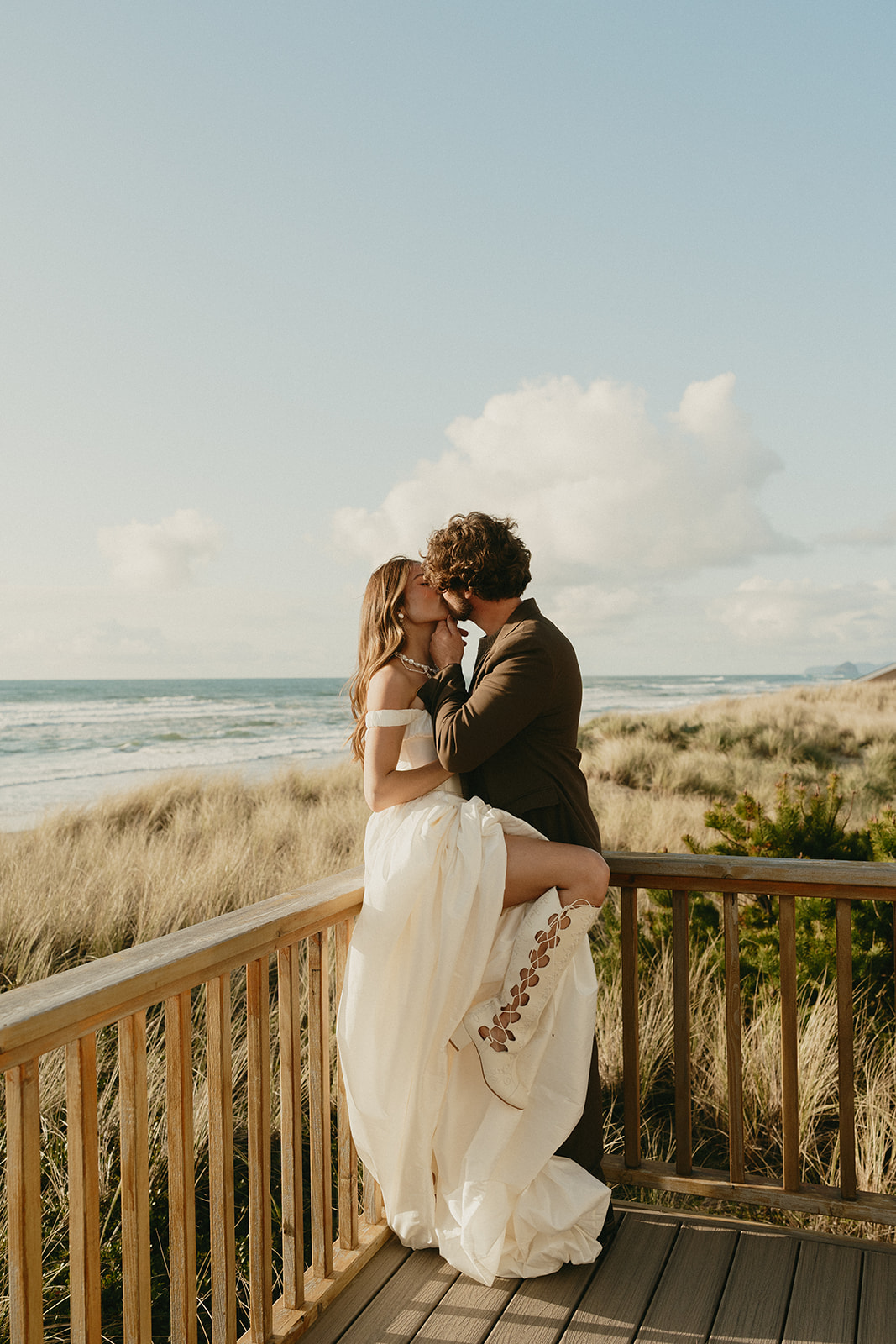With waves in the distance, the couple shares a kiss on the wooden deck during their Oregon Coast elopement, her boots peeking out from under her dress.