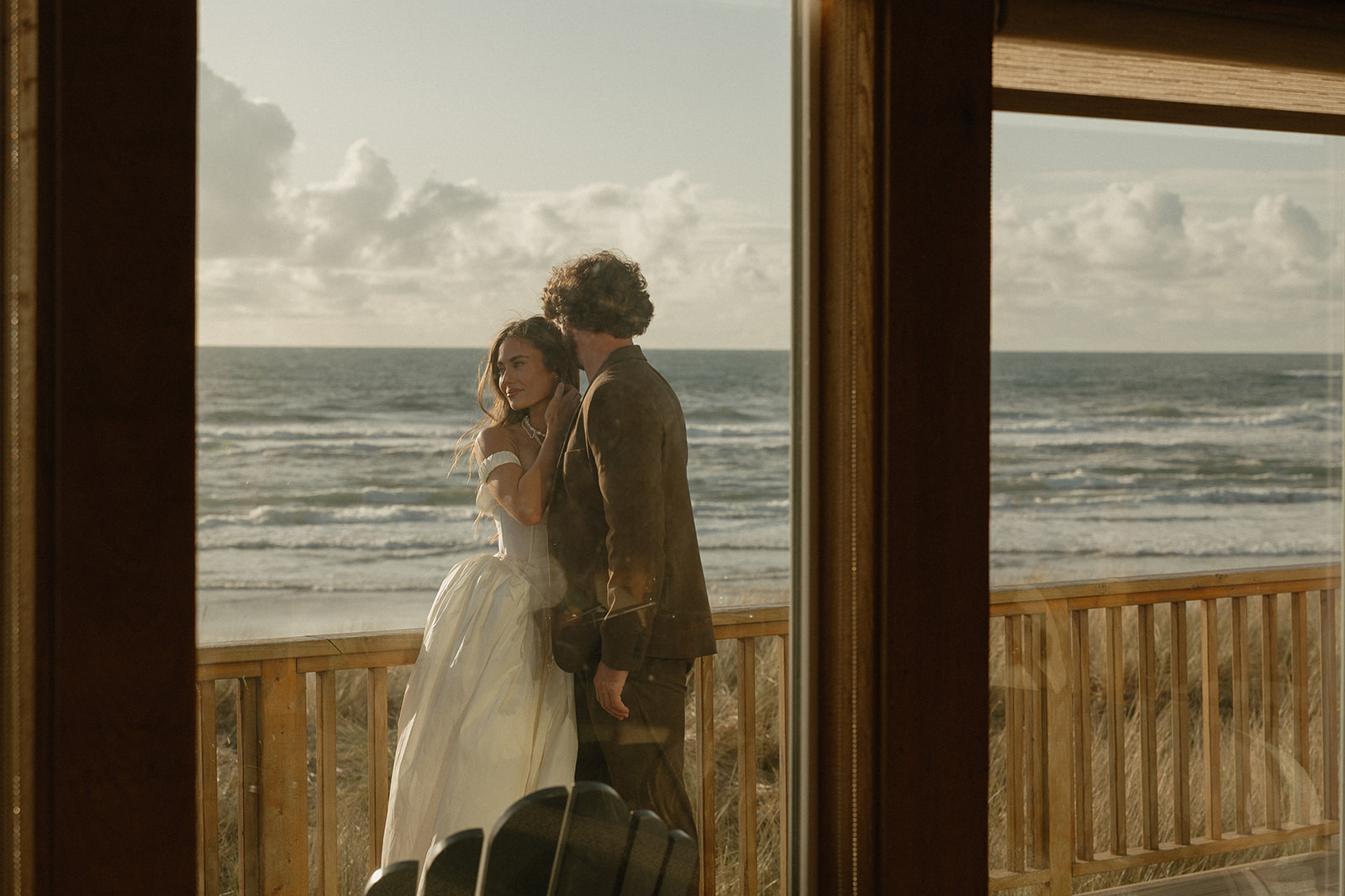 Seen through the glass, the couple stands wrapped in golden light, gazing out at the ocean during their peaceful Oregon Coast elopement.