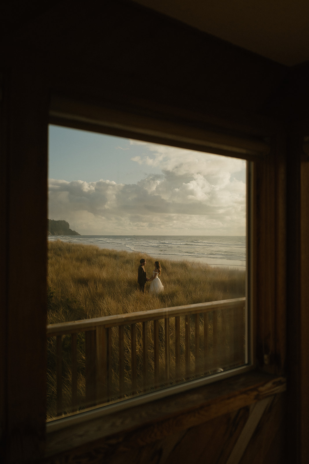 Romantic view through a window of a couple holding hands in the tall coastal grass during an Oregon Coast elopement.