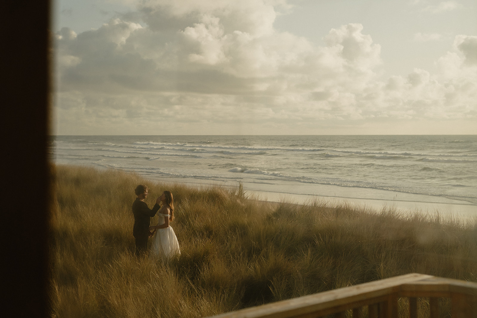 Seen through a window, the couple shares a quiet moment in the dune grass during their Oregon Coast elopement, the shoreline stretching peacefully behind them.