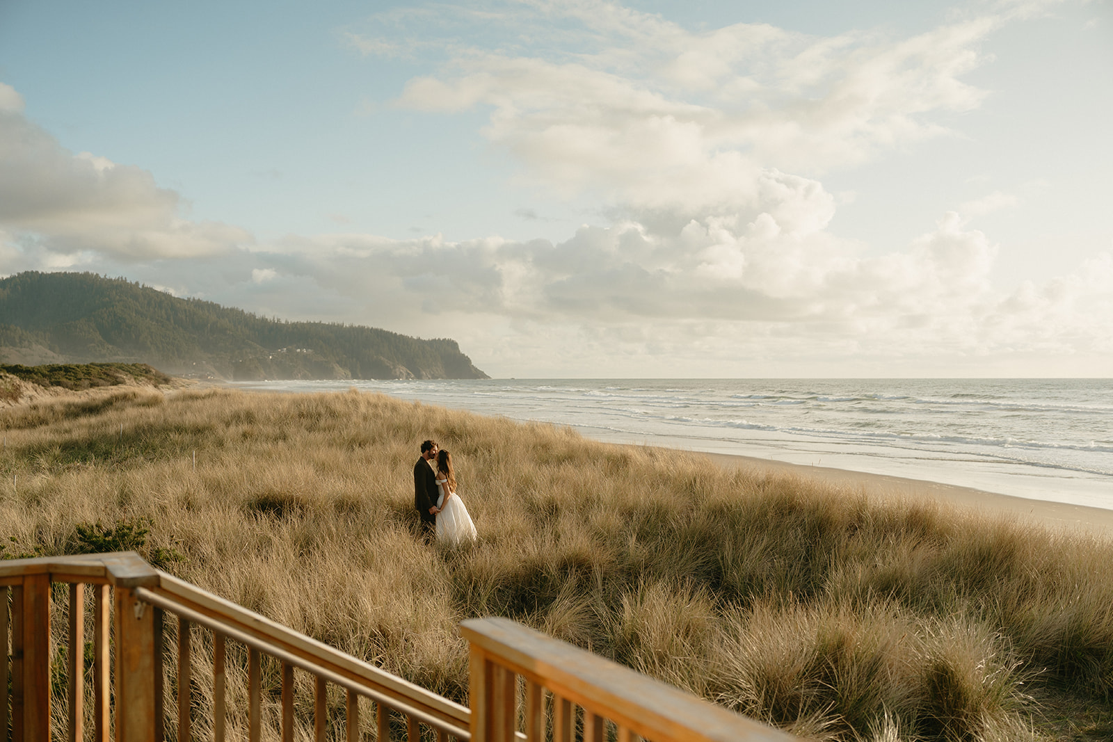 A couple embraces in the tall golden grass along the shoreline during their Oregon Coast elopement, with misty cliffs rising in the distance under soft morning light.