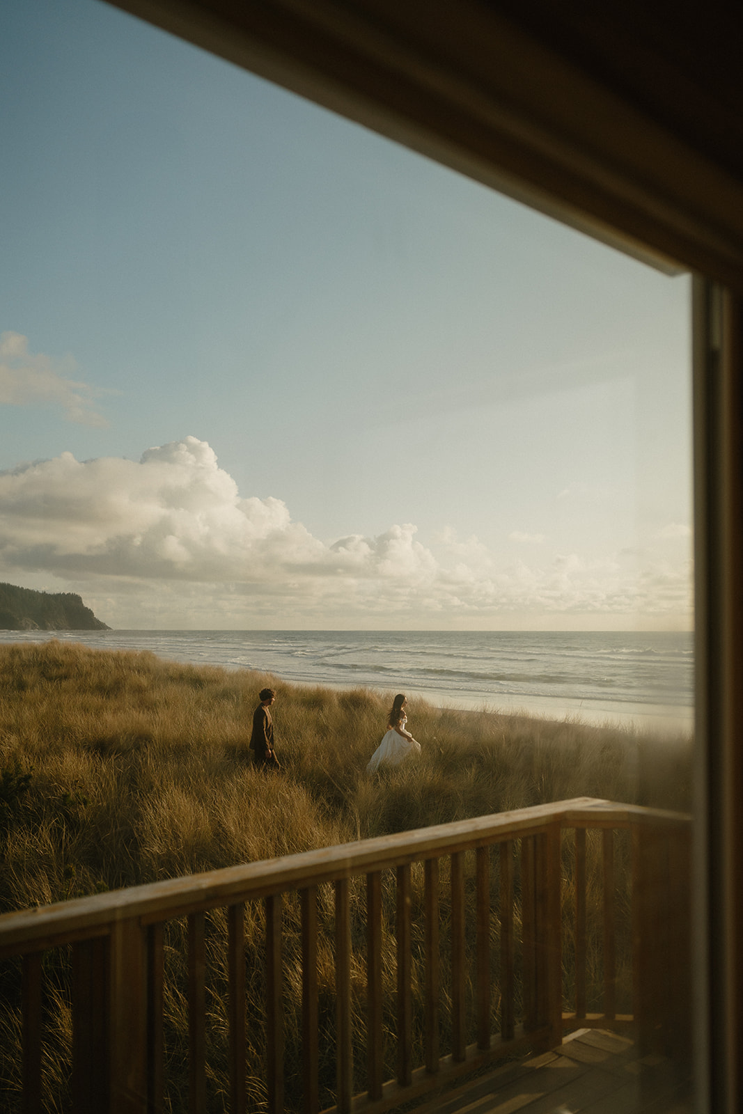 A couple walks through windswept grass during their Oregon Coast elopement, captured through the window of a quiet beachfront home as golden light softens the shoreline.