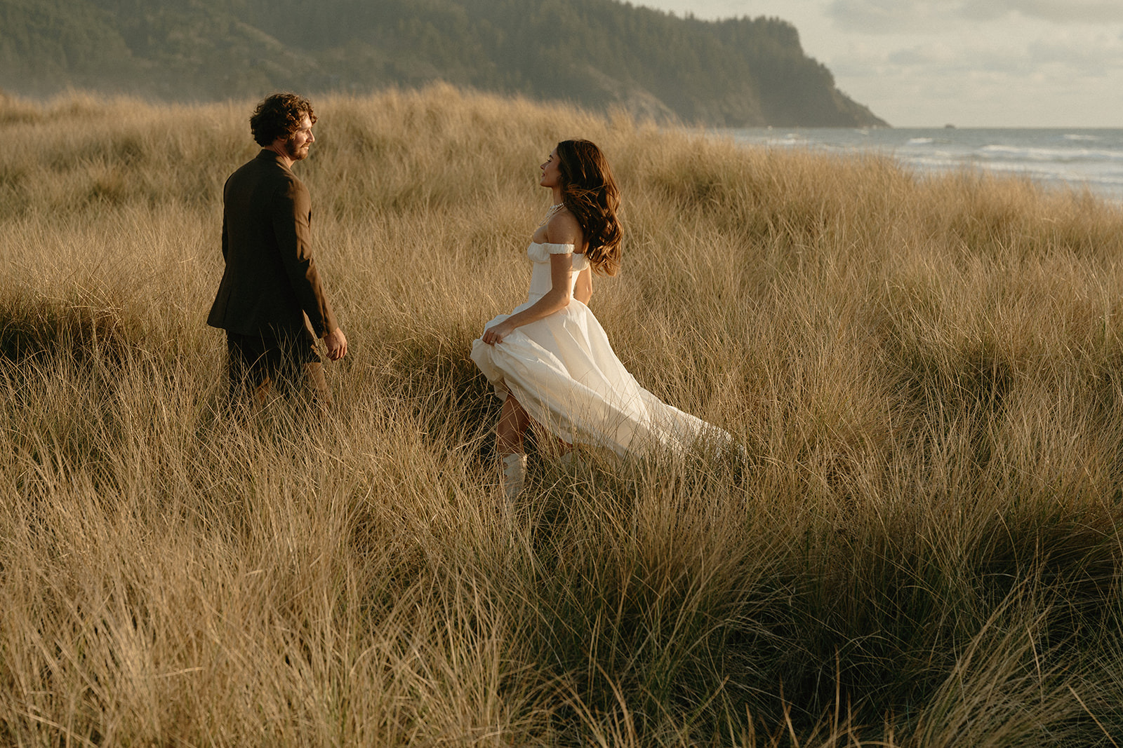 Bride in a flowing dress walks through coastal grass as her partner watches her lovingly during their Oregon Coast elopement.