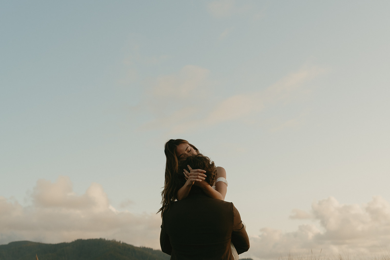 A bride wraps her arms around her groom’s neck as he lifts her gently, framed by the pastel sky and coastal hills at sunset.