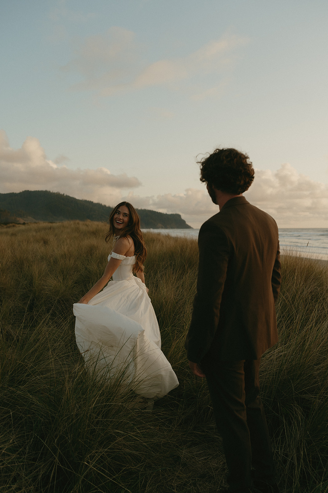 The bride turns back with a smile as she walks through coastal grasses during their Oregon Coast elopement, the groom following just behind under a glowing sky.