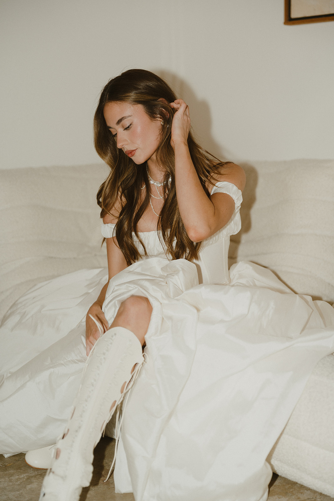 The bride sits thoughtfully on a cream-colored sofa, her off-the-shoulder gown and laced boots capturing the soft, editorial feel of an Oregon Coast elopement morning.