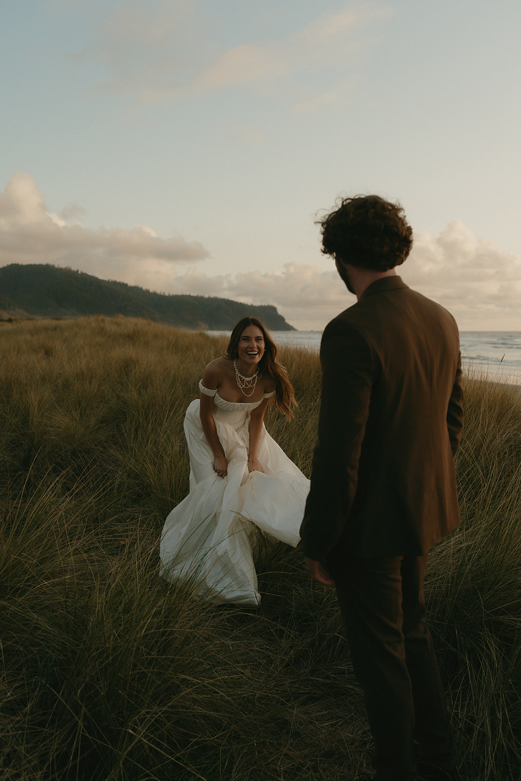 Bride laughing and holding her dress as she walks through tall beach grass toward her partner during an Oregon Coast elopement.