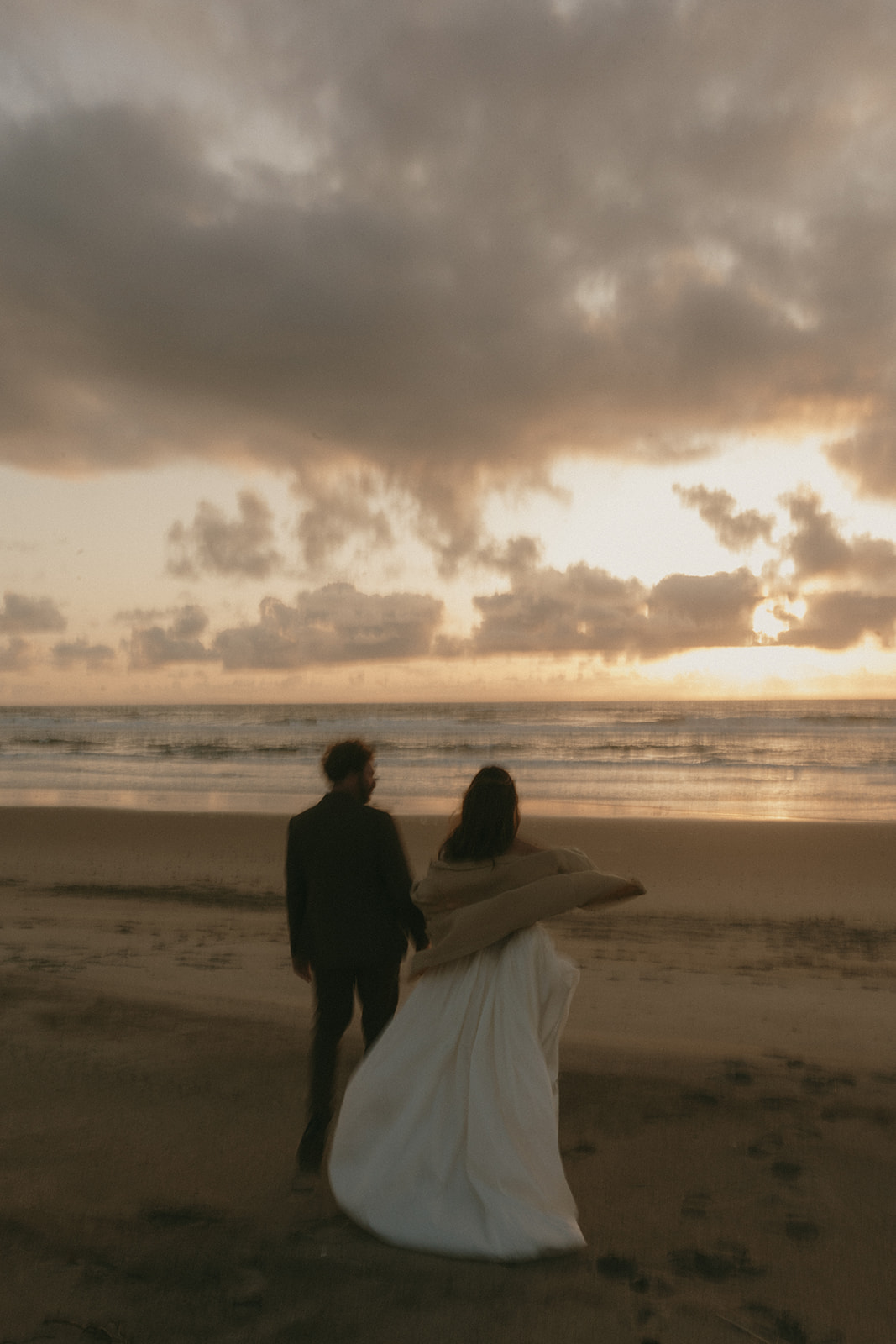 A couple walks along the shoreline during their Oregon Coast elopement, the bride’s dress trailing in the sand as the sky glows with soft evening light.