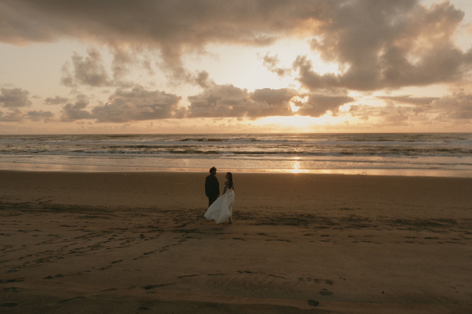 Wide shot of couple walking along the shoreline at sunset during their Oregon Coast elopement, silhouetted by the glowing sky.
