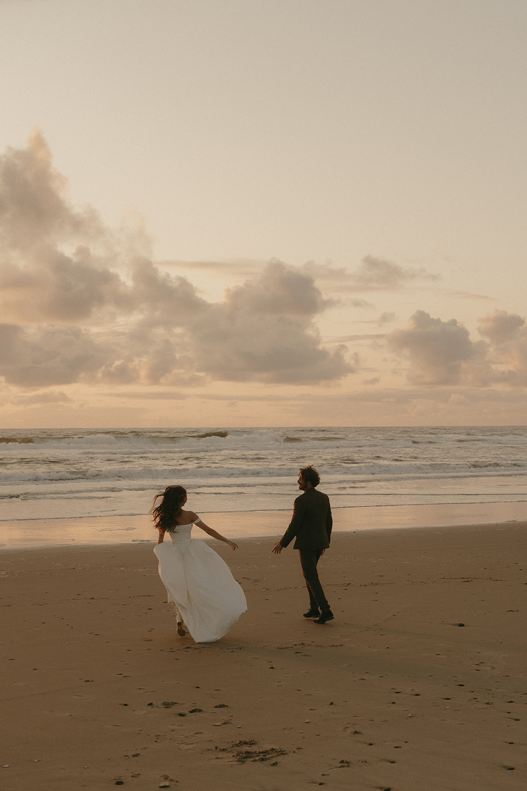 The couple runs barefoot along the shoreline during their Oregon Coast elopement, the bride’s dress flowing behind her in the golden light of sunset.