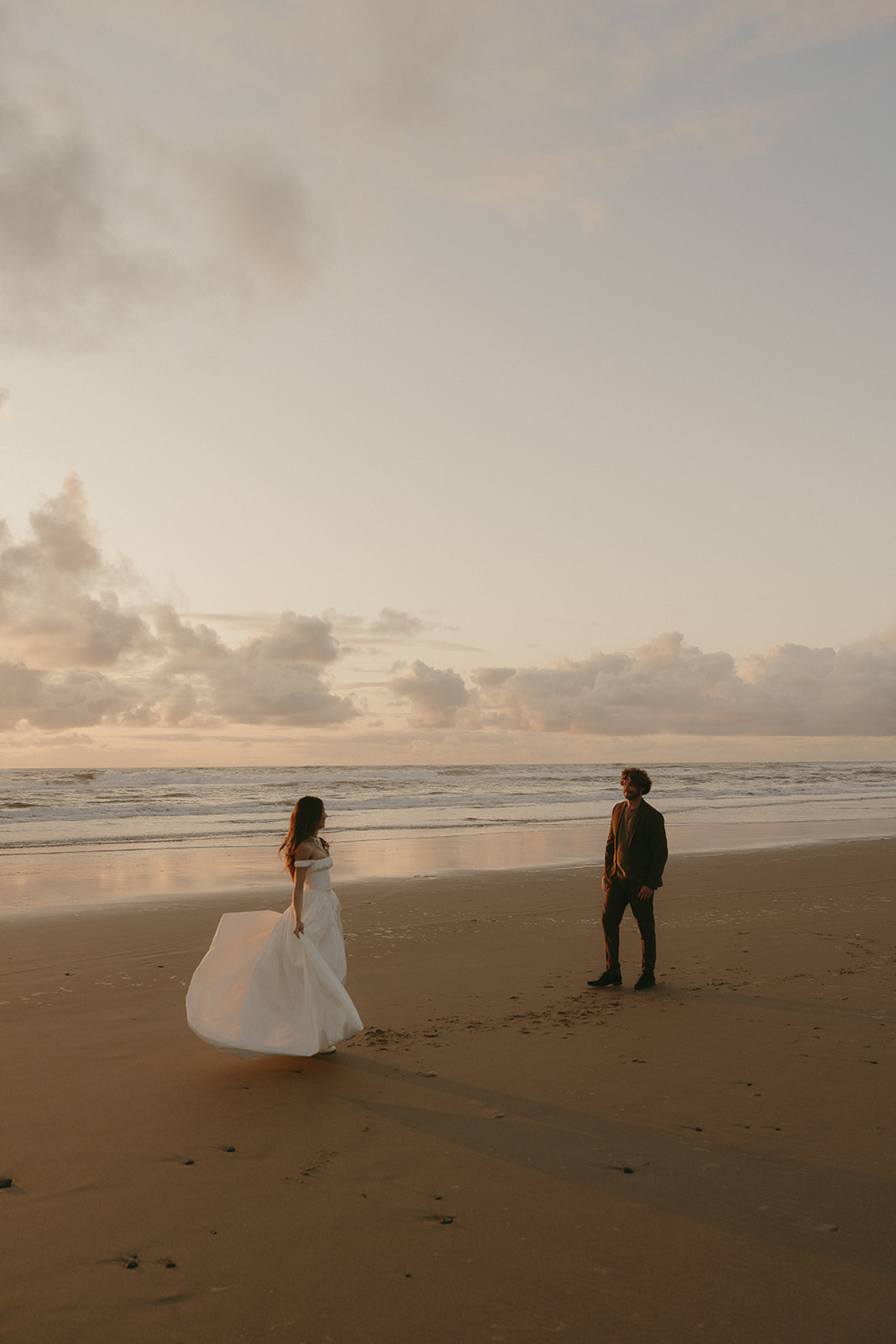 With soft light catching the hem of her dress, the bride steps across the sand toward her groom, the ocean stretching out under the pastel evening sky.