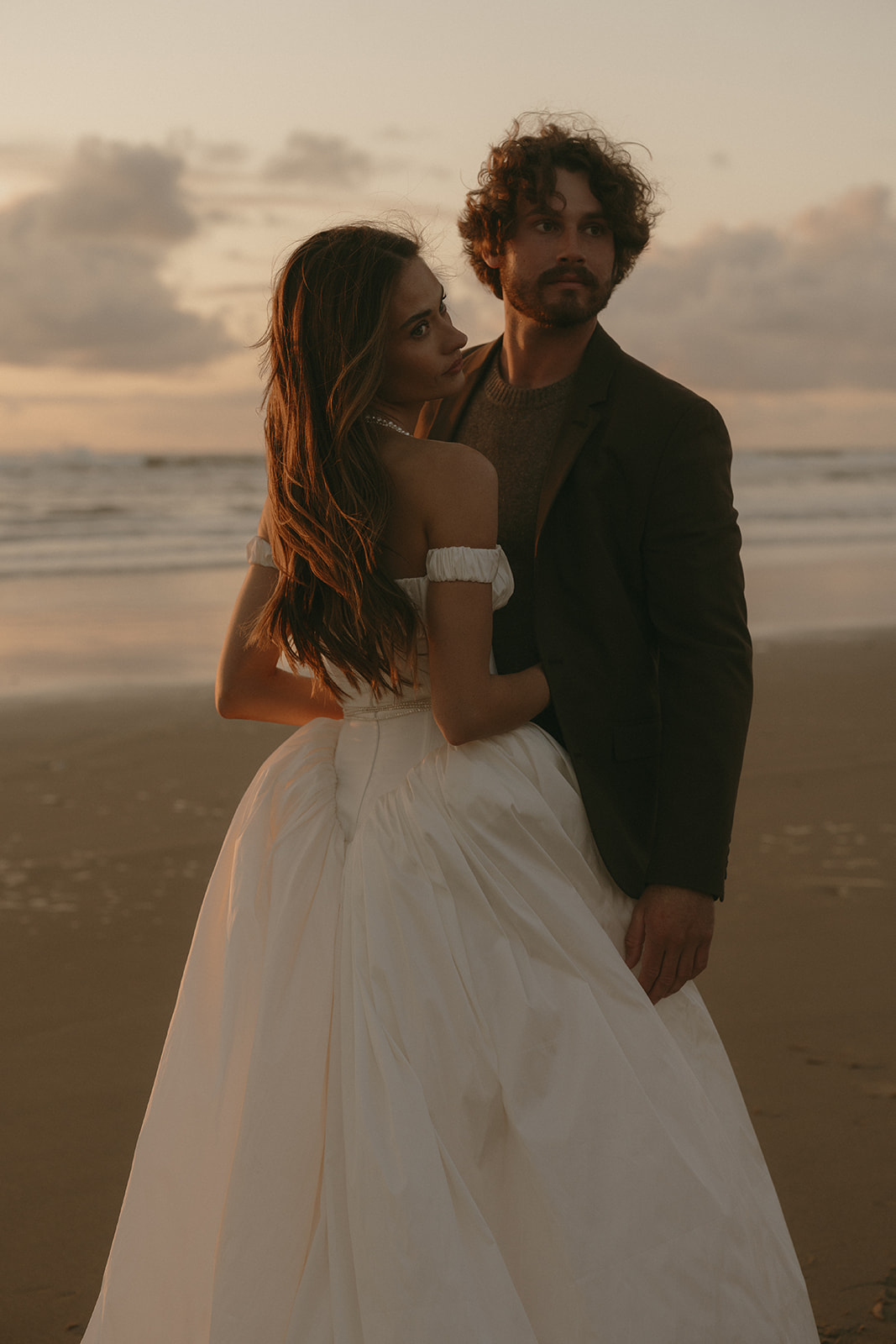 The bride rests gently against her groom as they stand in the last light of day, both facing opposite directions but fully at ease.