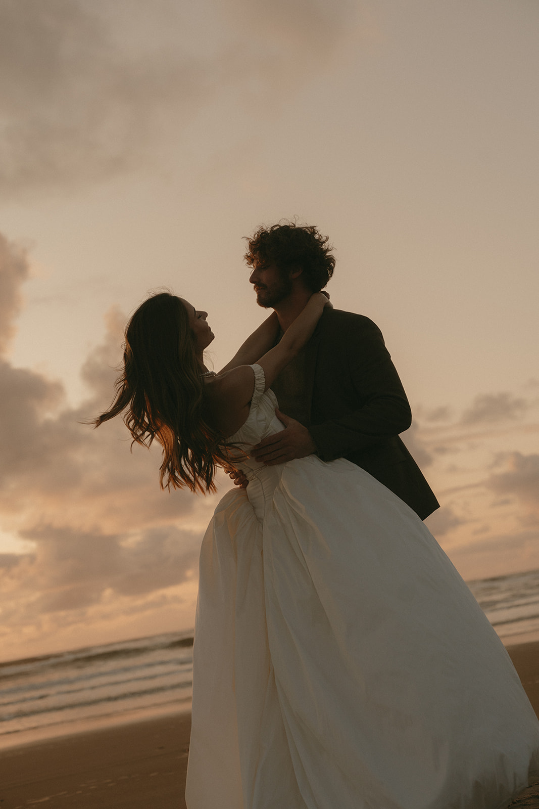 The groom holds the bride close on the beach at sunset during their Oregon Coast elopement, her dress catching the golden wind as they laugh together.