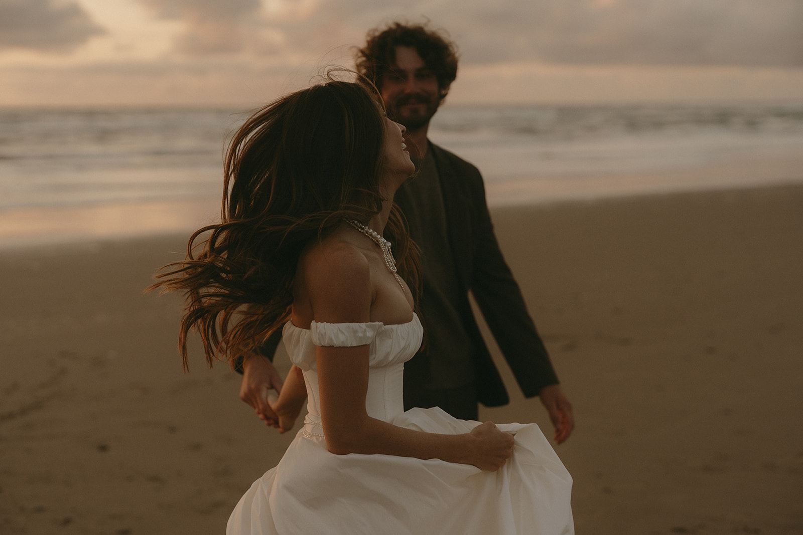 Captured in motion, the bride laughs as she runs hand-in-hand with her partner along the beach, her gown catching the golden hour breeze.