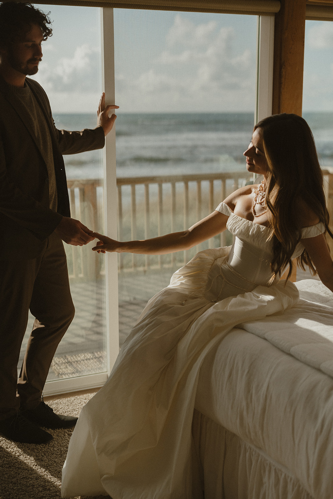 Soft window light pours in as the couple shares an intimate moment indoors, hands reaching across the quiet space between them.