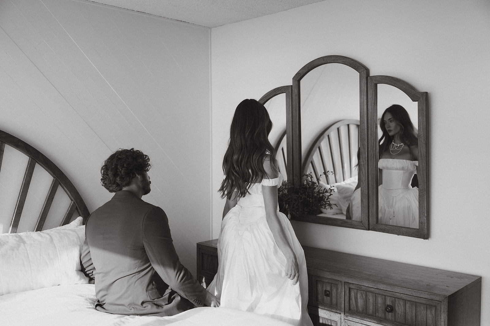 A cinematic black-and-white portrait of the bride reflected in the mirror as her groom watches from the bed—stillness and anticipation suspended between them.
