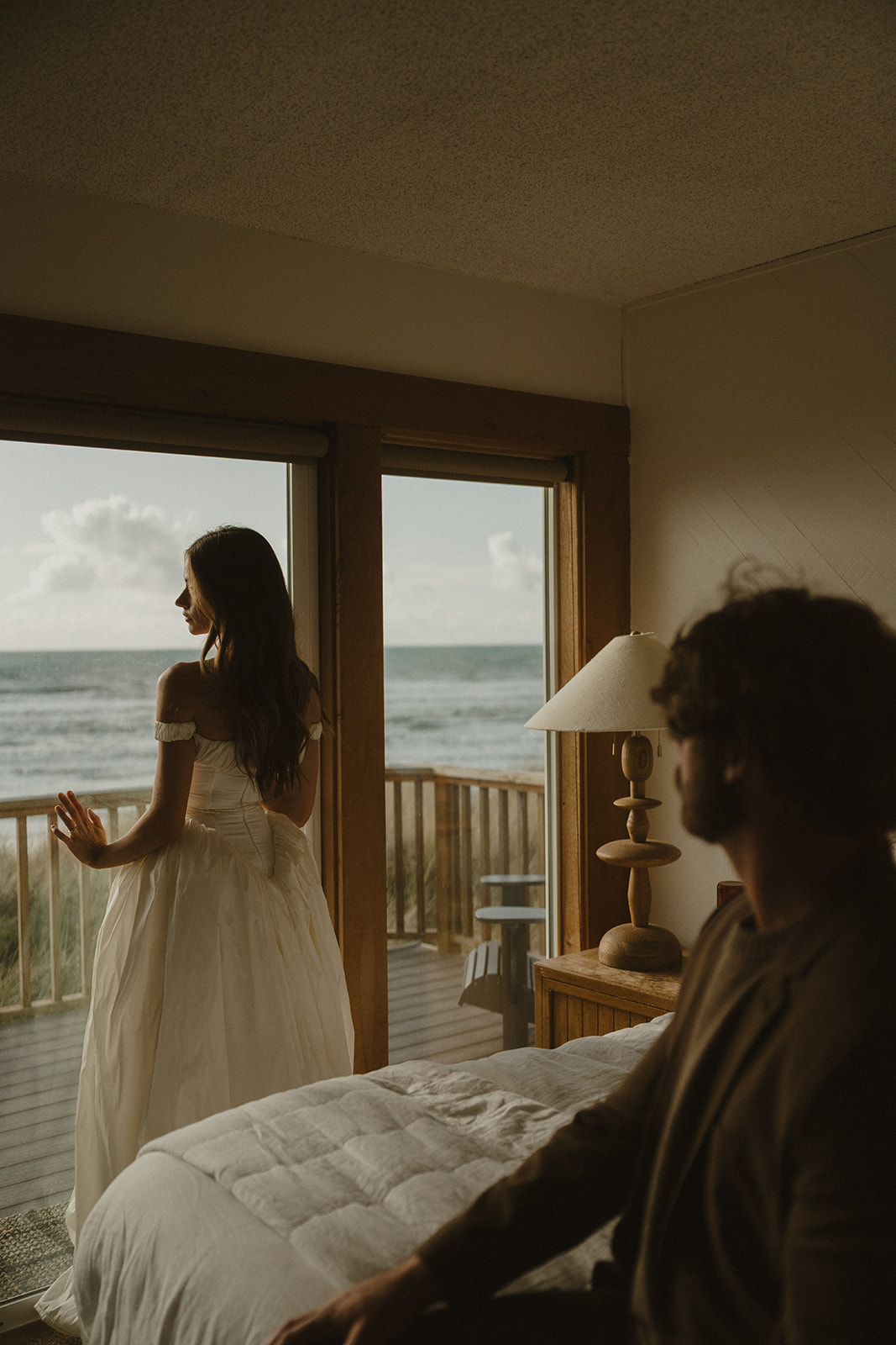 The bride gazes out the window, hands resting lightly on the glass, while her partner sits behind her on the bed, bathed in soft morning light.