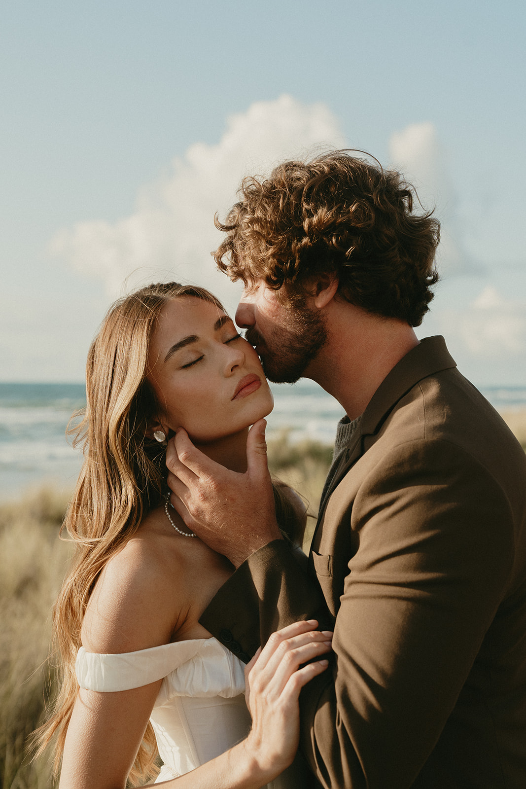 With wind catching her hair, the bride leans into her partner’s embrace as he presses a kiss to her forehead during their Oregon Coast elopement.